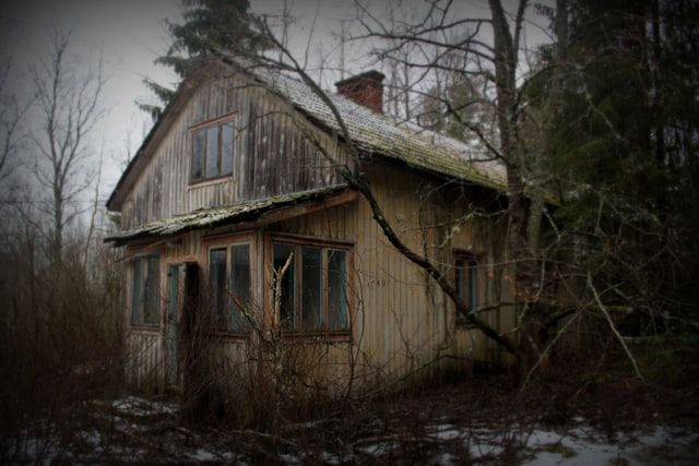 A photo of a house that looks abandoned, and possibly burned out. The roof is green and falling down, parts of the outside walls are blackened and the trees around the house look dead and have no leaves. A light smatter of snow is on the ground. 