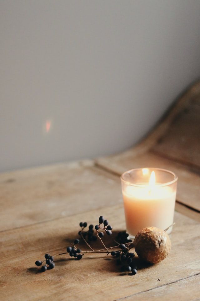 A candle sits on a wooden table. A lens flair shows as a spot of reddish light on the beige wall from the flame. Dried herbs sit in front of the candle. 