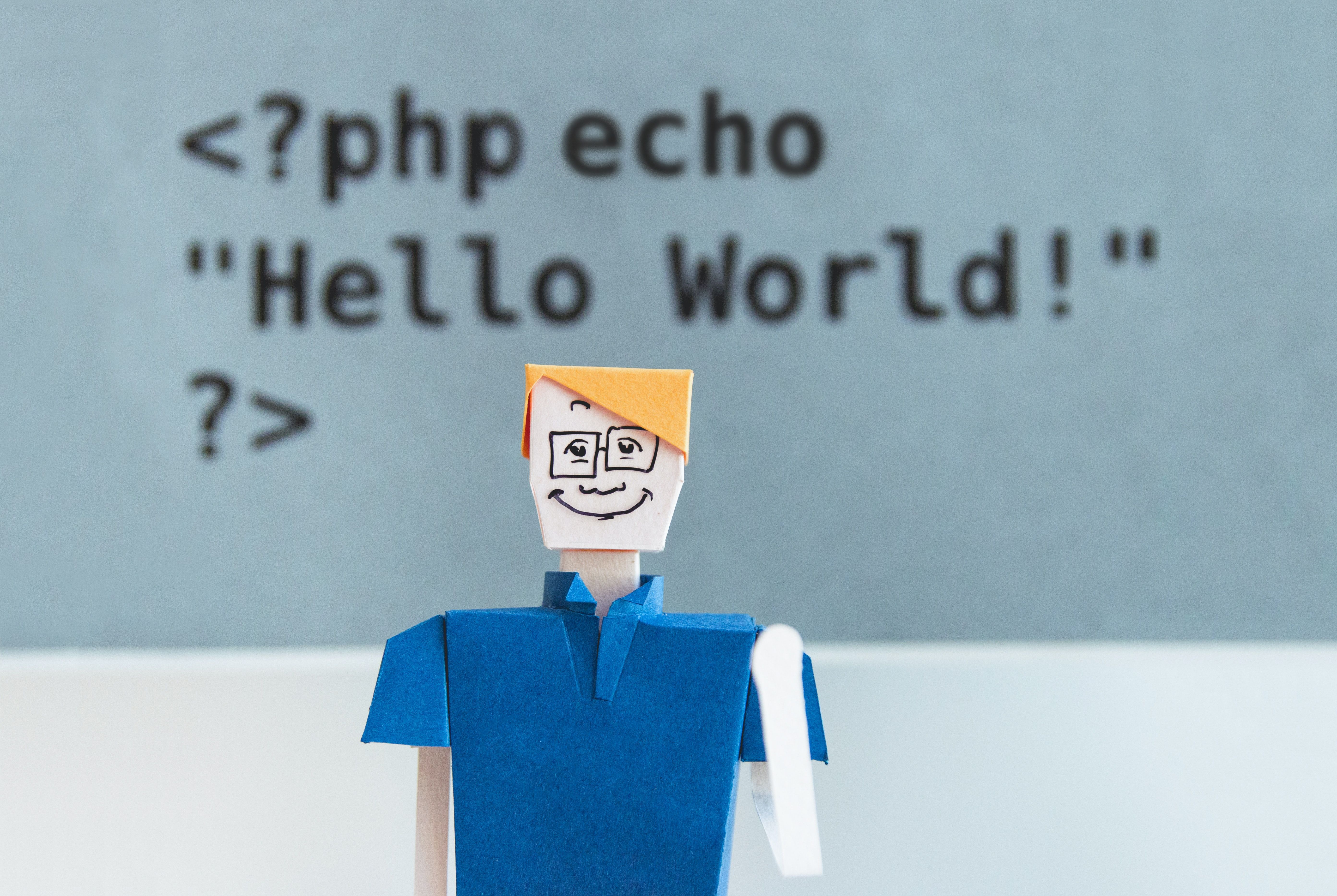 Photo of a paper doll man wearing a blue shirt smiling almost reluctantly at the camera, with PHP code written on the wall behind him