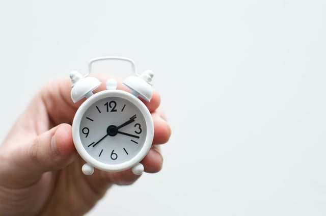 White background with a hand holding a small white alarm clock showing 2:19