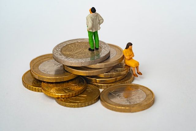 A small stack of gold coins and two tiny people on them. One is standing, looking to the sky and the other is sitting on a coin looking down with her arms crossed. 