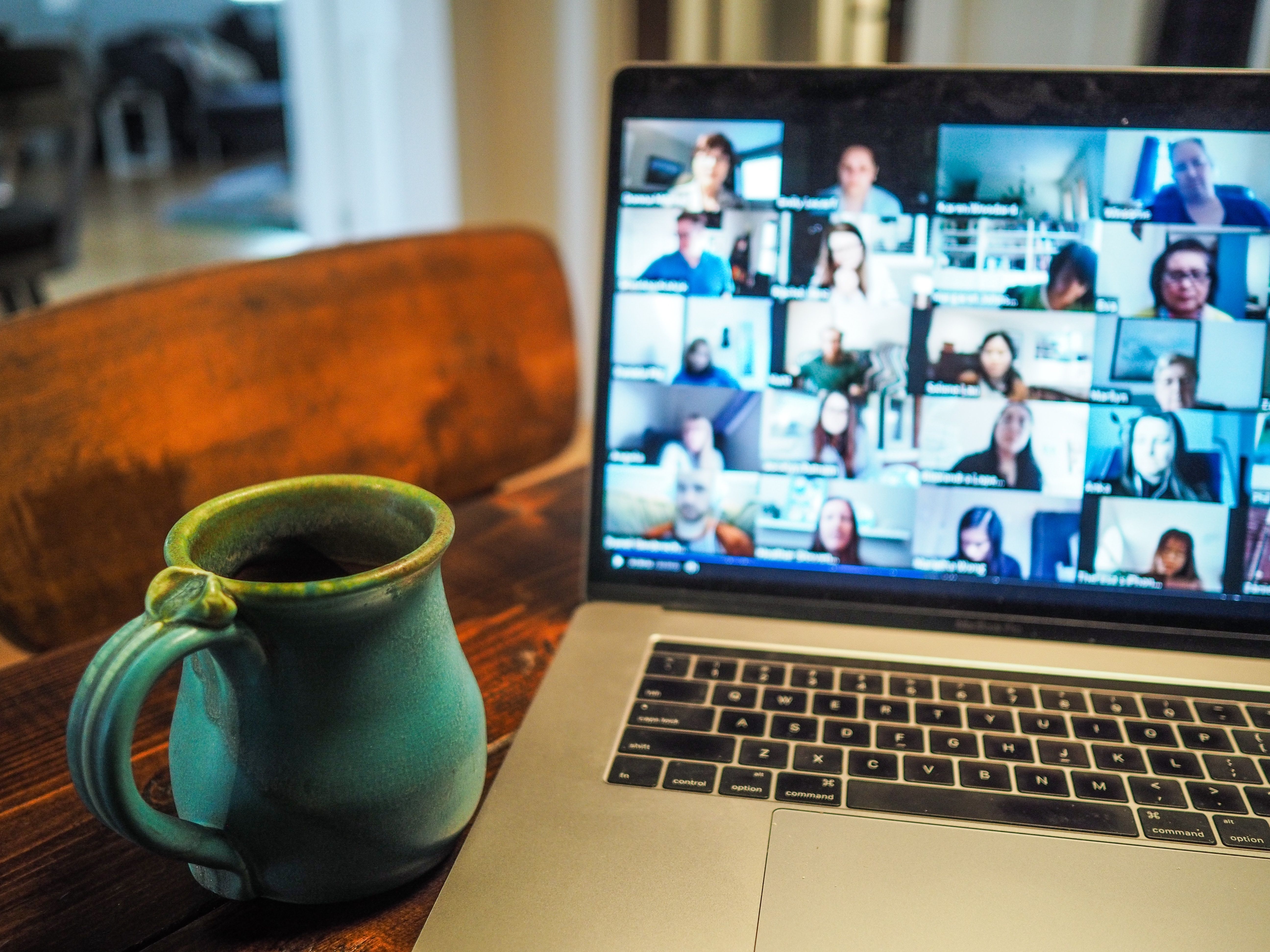 An image of a blue coffee cup on a table next to a laptop. On the laptop screen is 20 plus tiny images of people in a video conference meeting. 